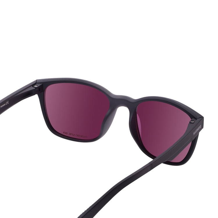 Black Shimano sport sunglasses with a matte black full-rim frame and mirrored bronze-gold lenses, shown from front and side angles on a light gray background. The rectangular lens shape features slightly rounded corners, with Shimano branding on the temple and textured temple tips for grip. The reflective lenses have a subtle pink-purple tint around the edges.