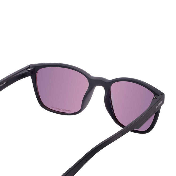 Matte black Shimano sunglasses with a lightweight full-rim frame and mirrored red-pink lenses, shown from front and side views on a light gray background. The rectangular lenses have gently rounded edges, while the slim black temples feature Shimano branding and textured temple tips for added grip. The reflective lenses display a vibrant rose-red finish with subtle purple tones around the lens edges.
